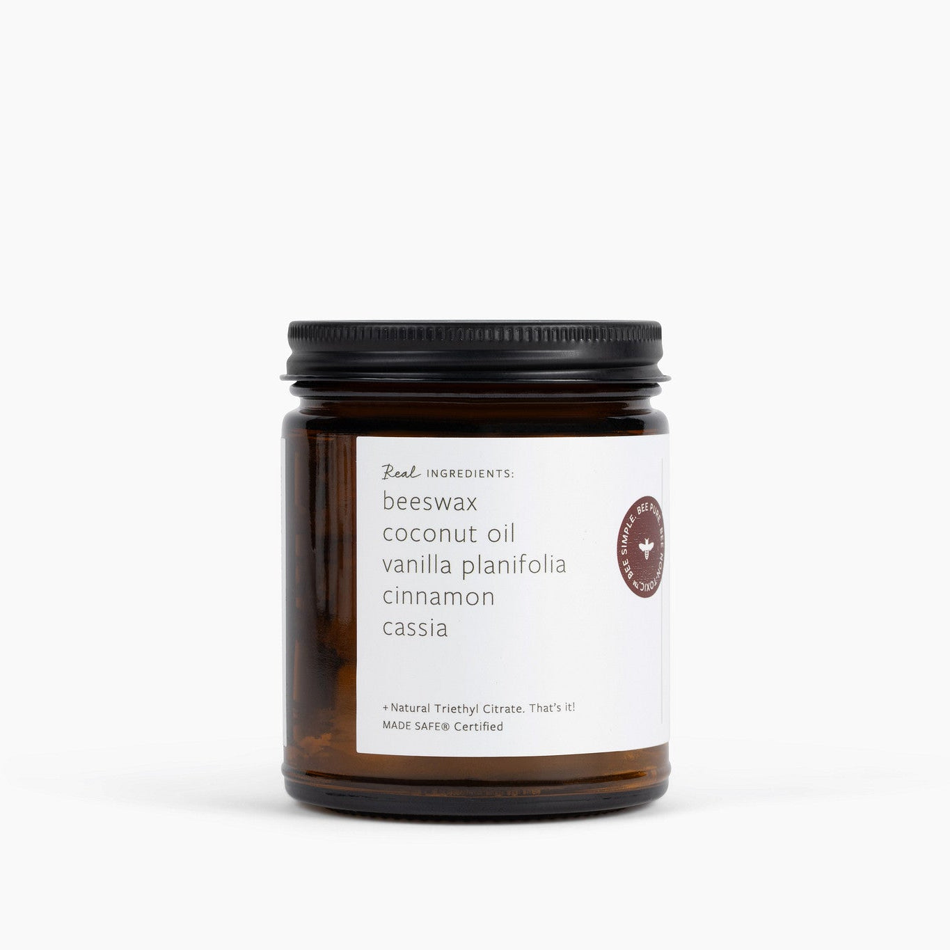 An amber glass jar candle with a black metal lid and a white label, photographed on a clean white background. The label lists ingredients including beeswax, coconut oil, vanilla planifolia, cinnamon, and cassia.