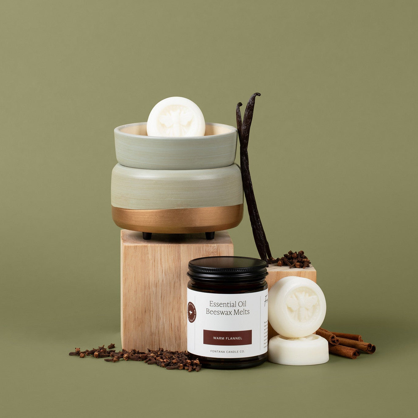 Wax melts and wax melter set to an olive green background, surrounded by vanilla beans, cloves and cinnamon sticks
