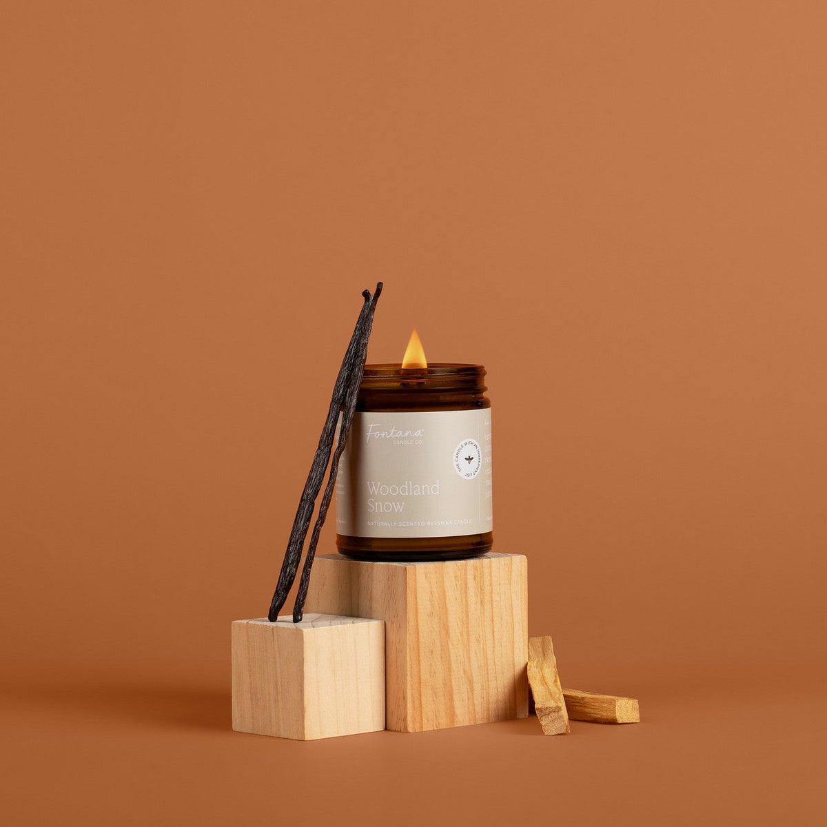 A lit Woodland Snow  candle in an amber glass jar with vanilla stick on wooden blocks against a brown background