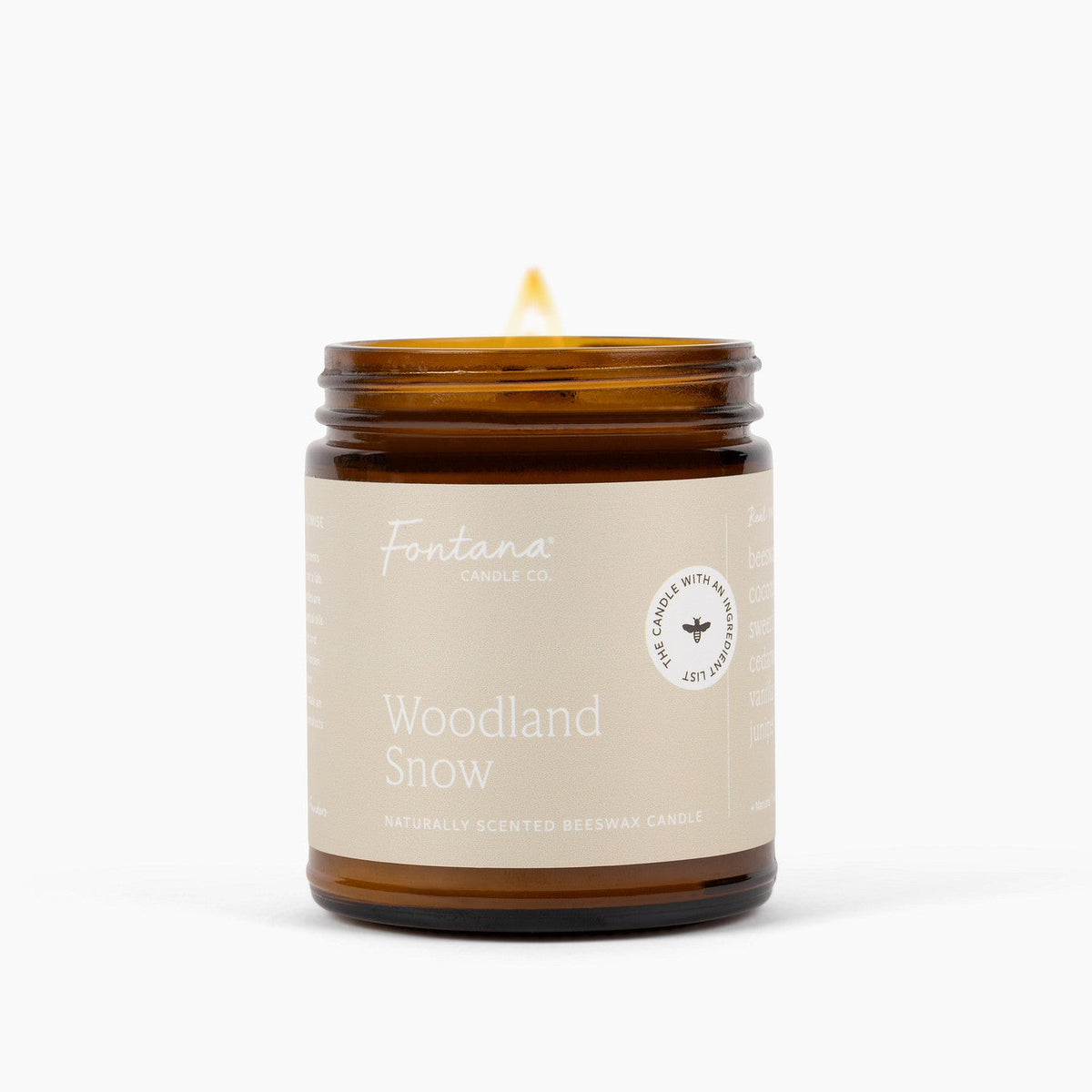 An amber glass jar candle with a lit wick, featuring a dark brown label that reads 'Fontana Candle Co. Woodland Snow' on a clean white background.