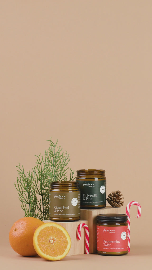 Three candles in glass jars with different scents on a brown background, surrounded by oranges and candy canes.