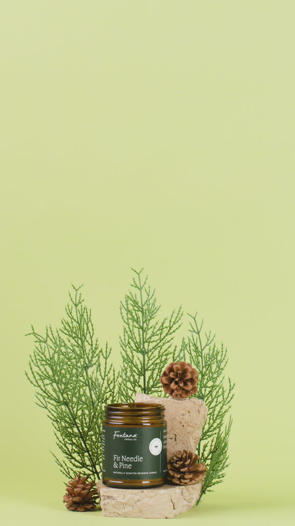 Candle with 'Fir Needle & Pine' label, surrounded by greenery and a hand placing a pine cone on top, against a light green background.