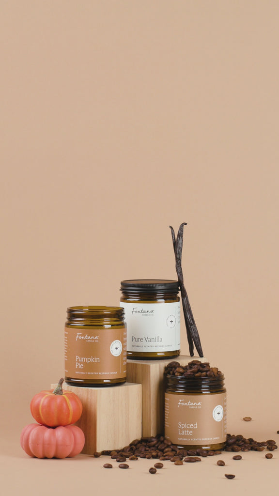 Candles with visible labels on a brown background, accompanied by pumpkins and coffee beans.