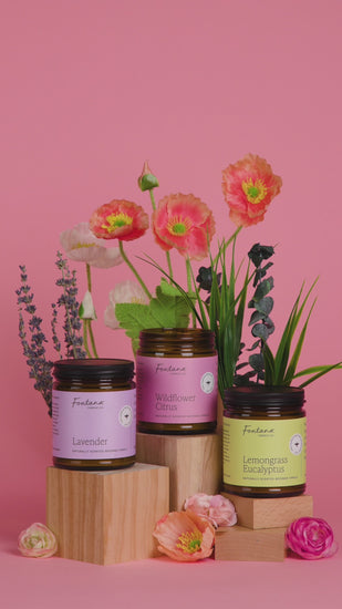 a video of someone lighting the wildflower citrus 9oz jar candle, with lemongrass eucalyptus and lavender next to it, on a pink background