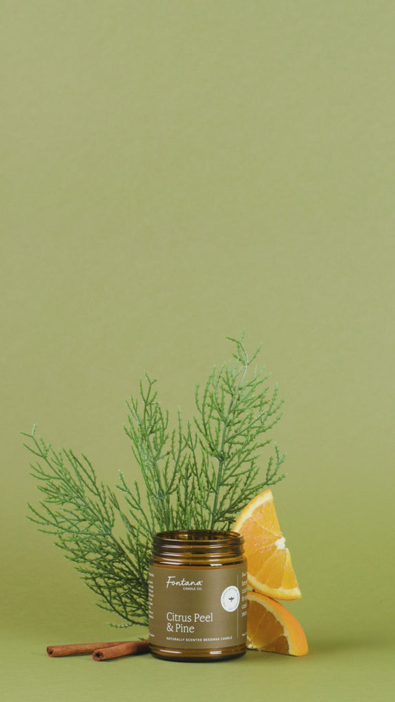 Candle with 'Citrus Peel & Pine' scent surrounded by orange slices, cinnamon sticks, and greenery on a green background.