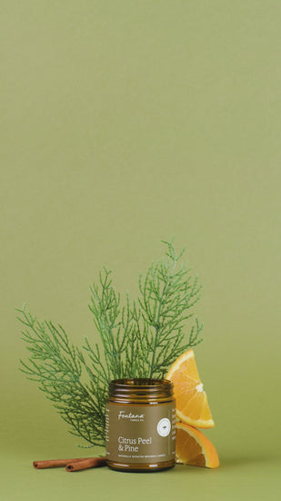 Candle with 'Citrus Peel & Pine' scent surrounded by orange slices, cinnamon sticks, and greenery on a green background.
