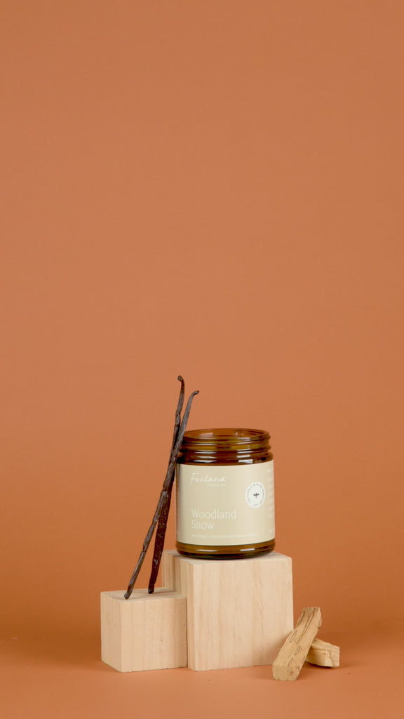Woodland Snow amber glass jar candle with vanilla stick on wooden blocks against a brown background