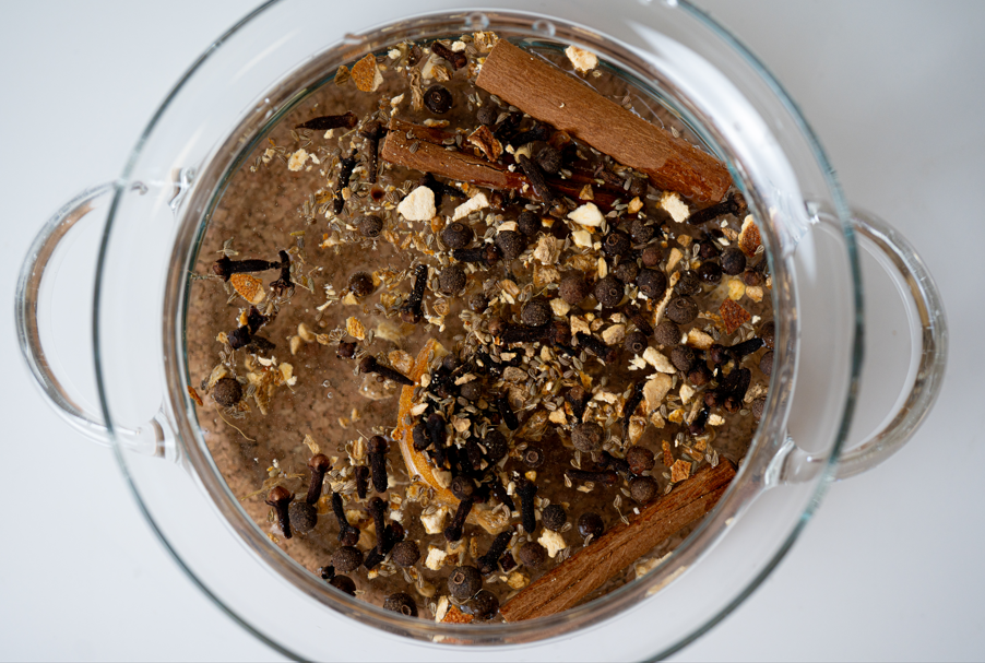 Glass pot with a mixture of ingredients inside including spices and cinnamon sticks.