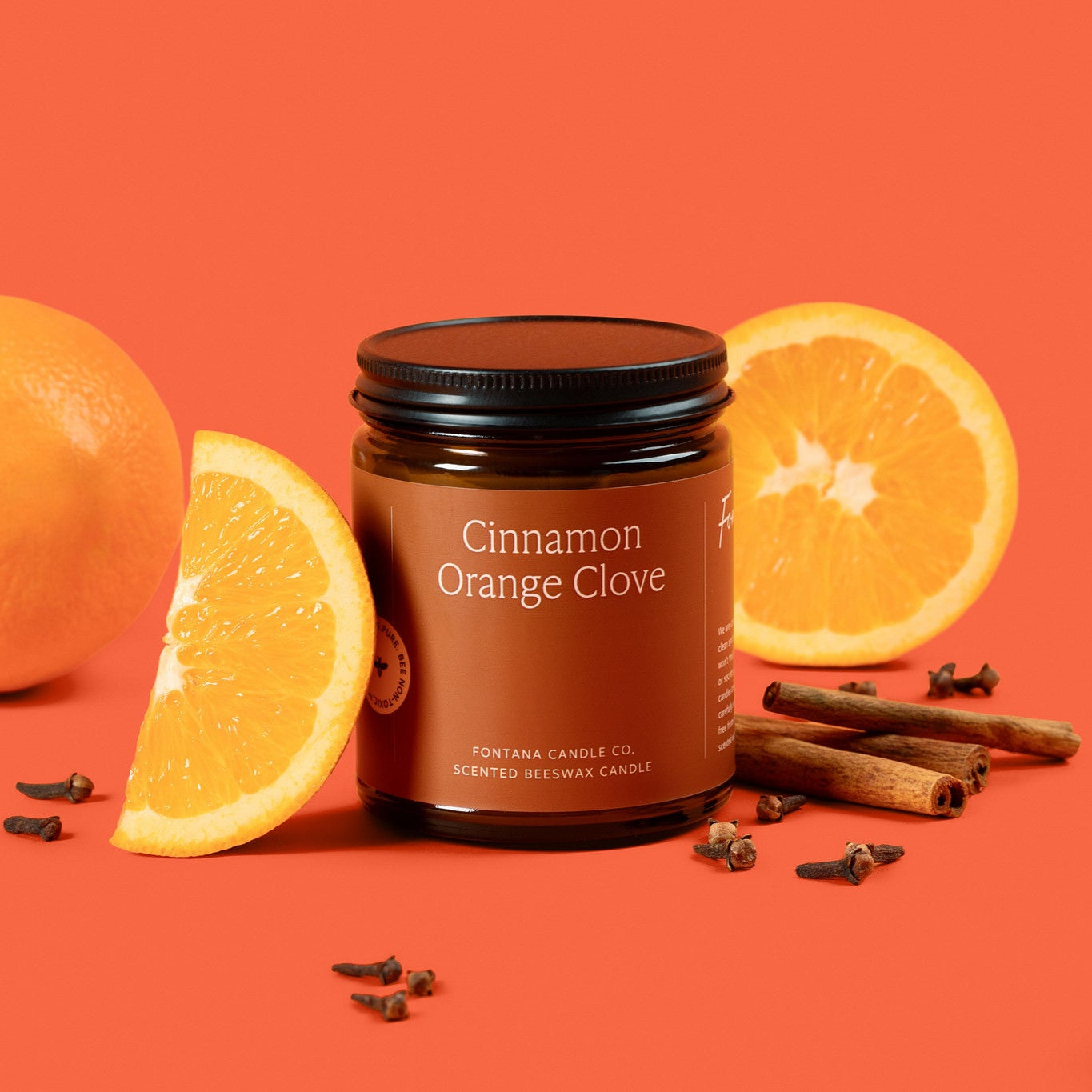 Amber glass jar candle with an orange label, surrounded by oranges, cinnamon sticks, and cloves. 