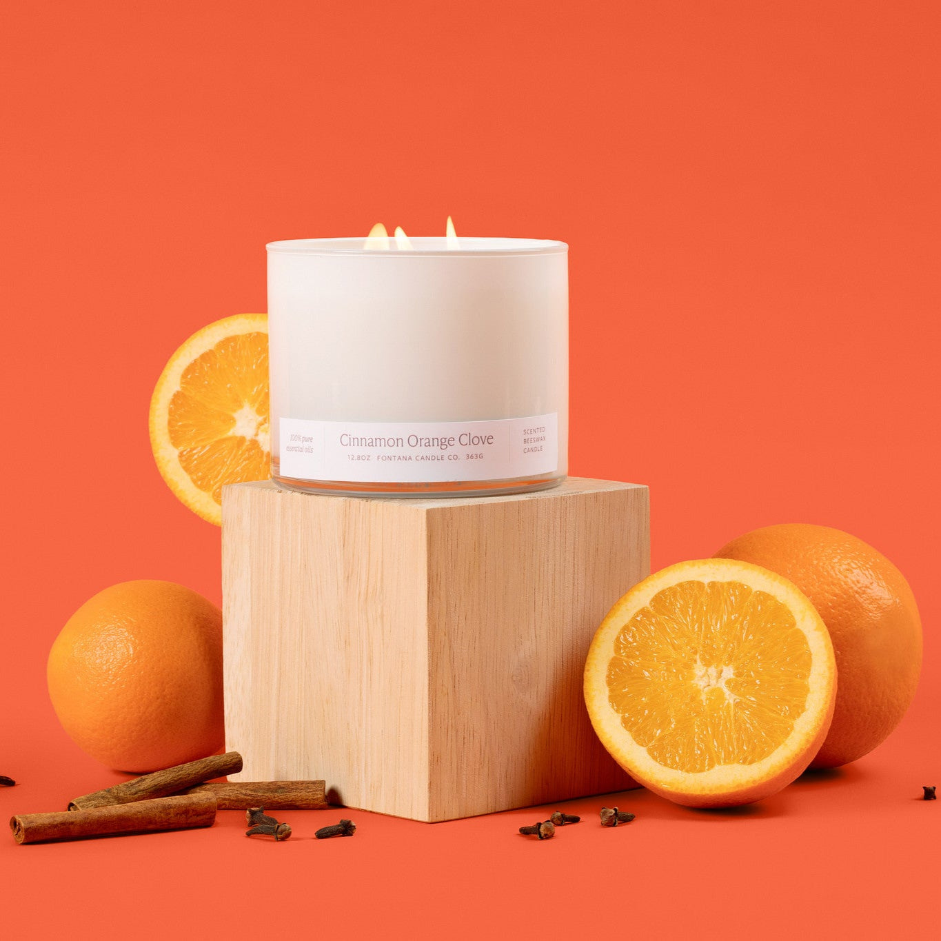 White glass 3-wick candle on a wooden block with oranges and cinnamon sticks on an orange background