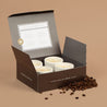 Open box containing 4 white ceramic espresso cups with on a brown background, surrounded by coffee beans.