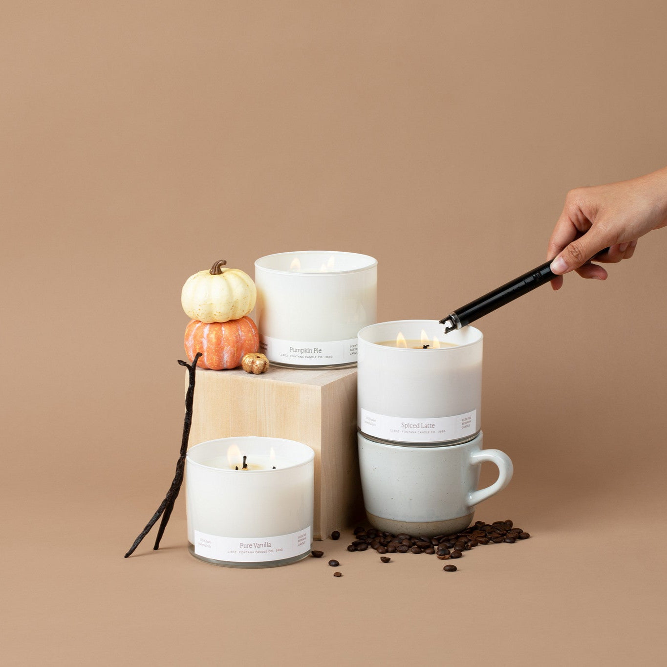 3 wick Spiced Latte Candle being lit with a black lighter, surrounded by vanilla beans, coffee beans, and small pumpkins on a beige background. Two other large 3 wick candles with white glass vessels are in the image: Pumpkin Pie, and Pure Vanilla.