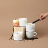 3 wick Spiced Latte Candle being lit with a black lighter, surrounded by vanilla beans, coffee beans, and small pumpkins on a beige background. Two other large 3 wick candles with white glass vessels are in the image: Pumpkin Pie, and Pure Vanilla.