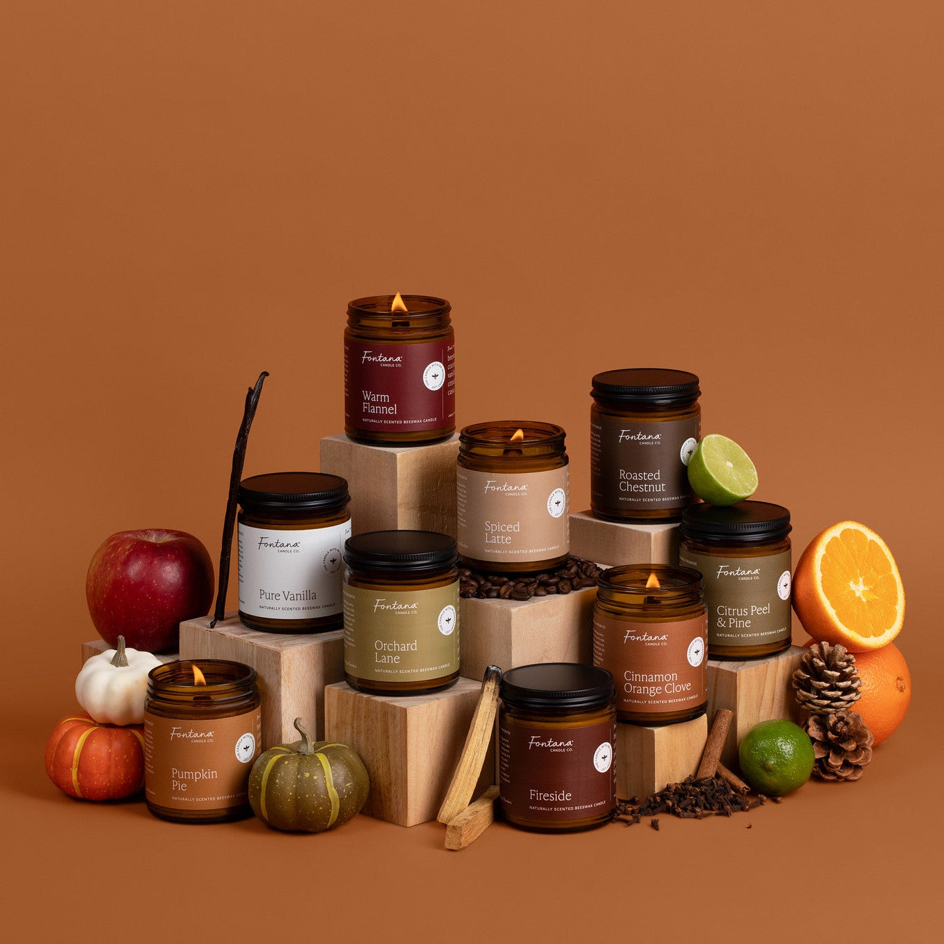 A styled product display of nine candles in amber glass jars with black lids, arranged on wooden blocks against a solid terracotta background. The candles are surrounded by autumn and citrus props including a red apple, a small pumpkin, a green lime, a cinnamon stick, pinecones, and an orange slice.