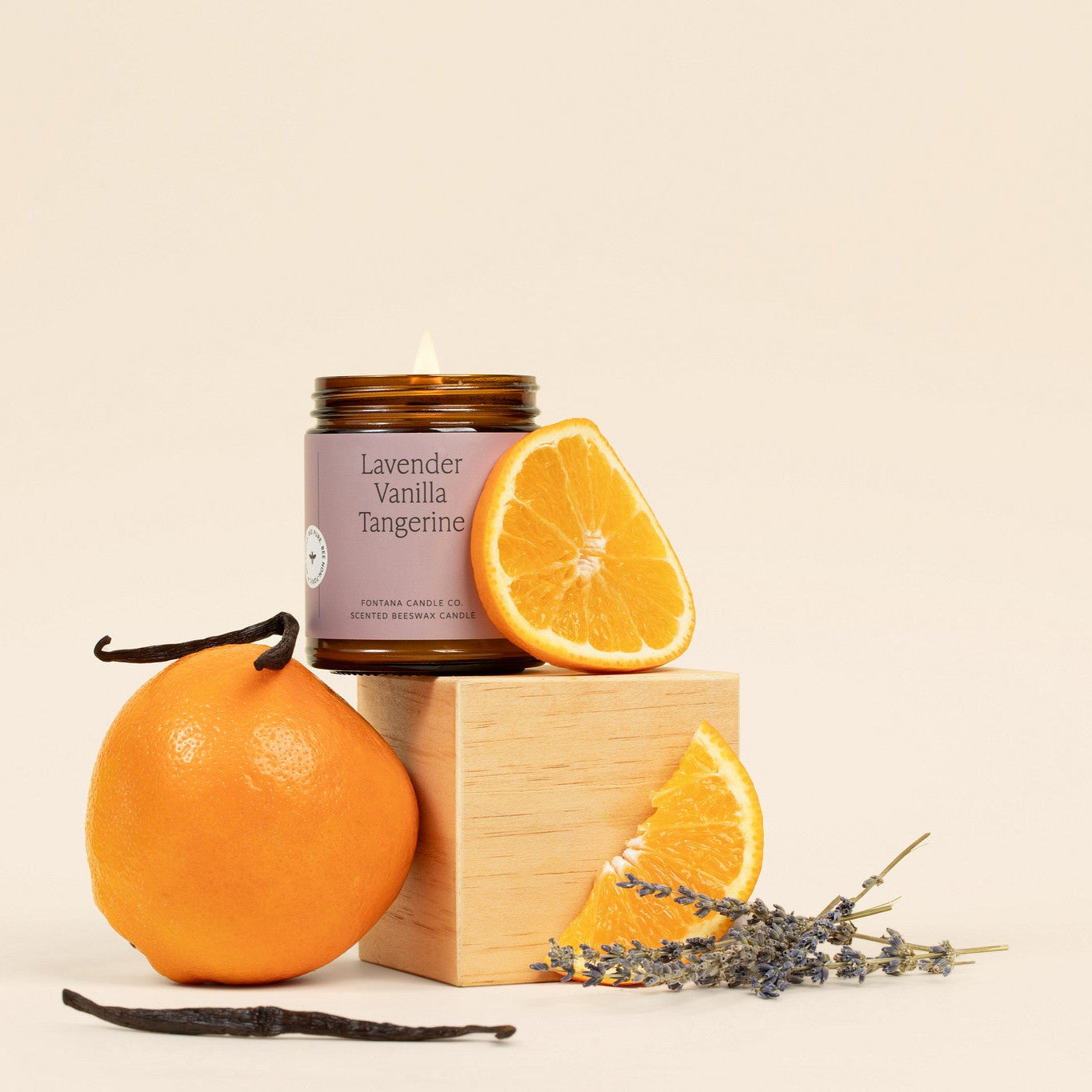 Lit amber glass jar candle with a light purple label, surrounded by oranges, lavender, and vanilla pods.