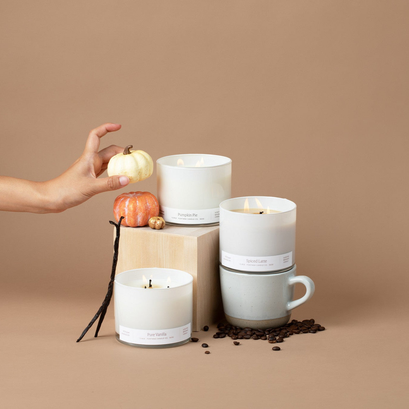 3 lit 3-wick candles in white glass vessels, surrounded by vanilla beans, coffee beans, and small pumpkins on a beige background. 