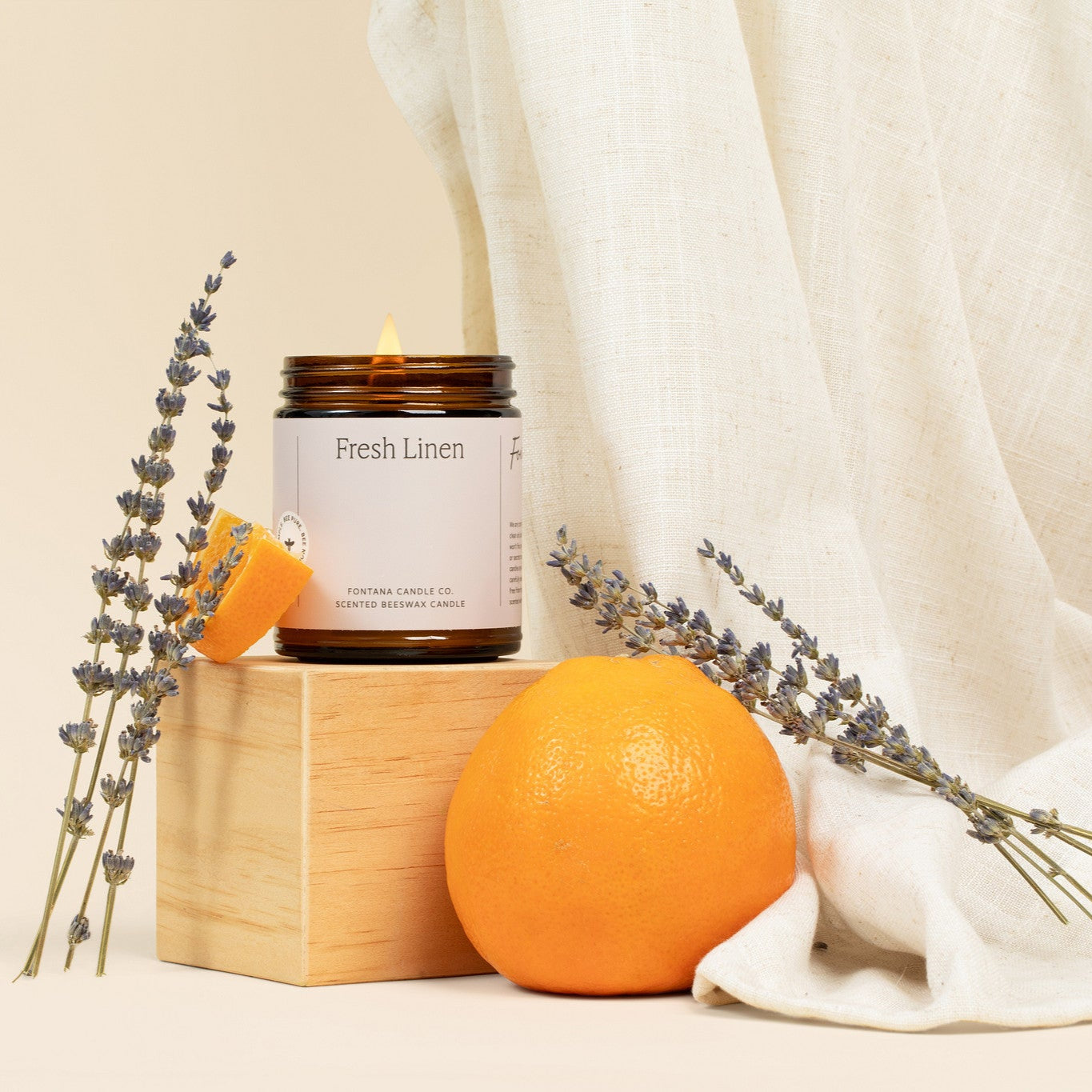 Lit amber glass jar candle with a light cream label, in front of linen sheets, lavender flowers, and oranges.
