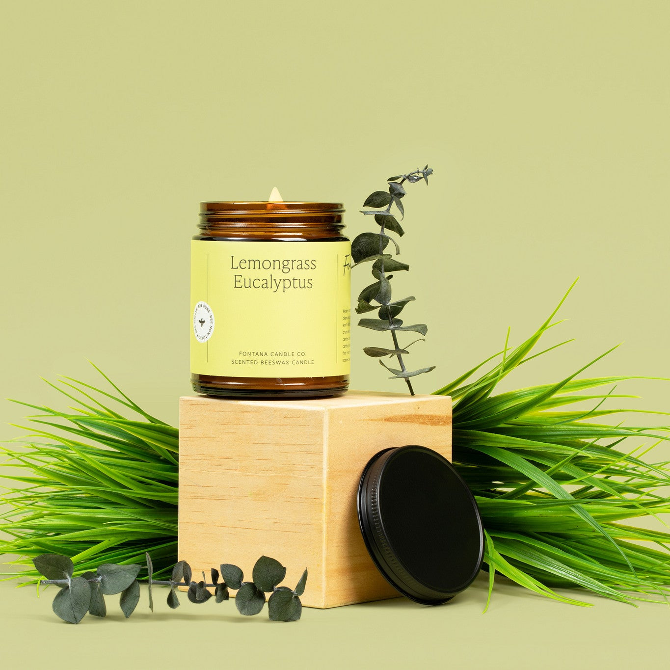Lit amber glass candle labeled 'Lemongrass Eucalyptus' on a wooden block with greenery on a light green background.