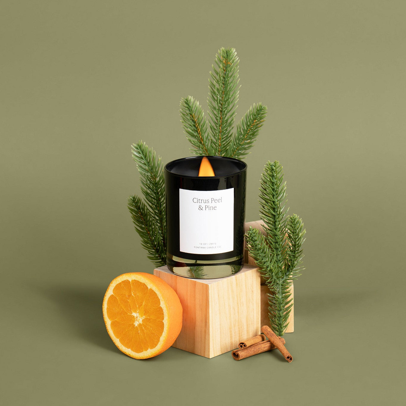 A black glass noir candle with a white label reading 'Citrus Peel &amp; Pine' sits on a light wood block, styled with green pine branches, a halved orange, and cinnamon sticks against a solid olive green background.