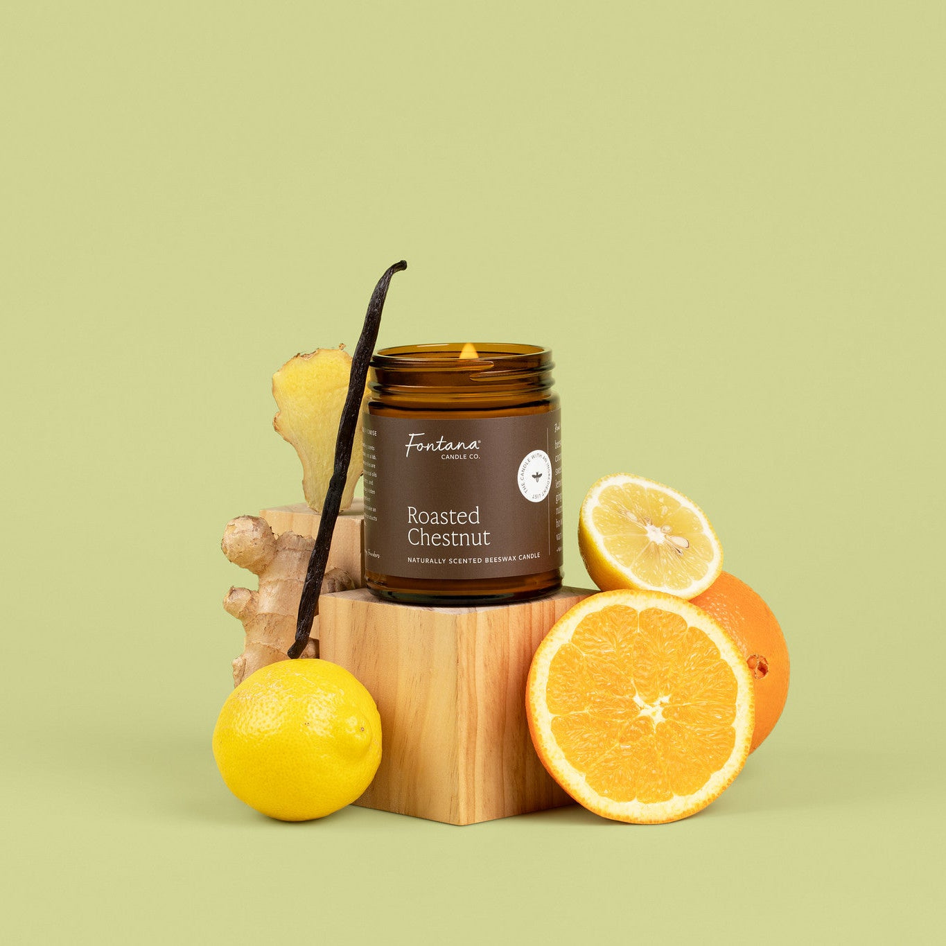 An amber glass jar candle labeled 'Roasted Chestnut' styled on a wooden block with a vanilla bean, ginger root, a whole lemon, and sliced oranges against a solid light olive-green background.