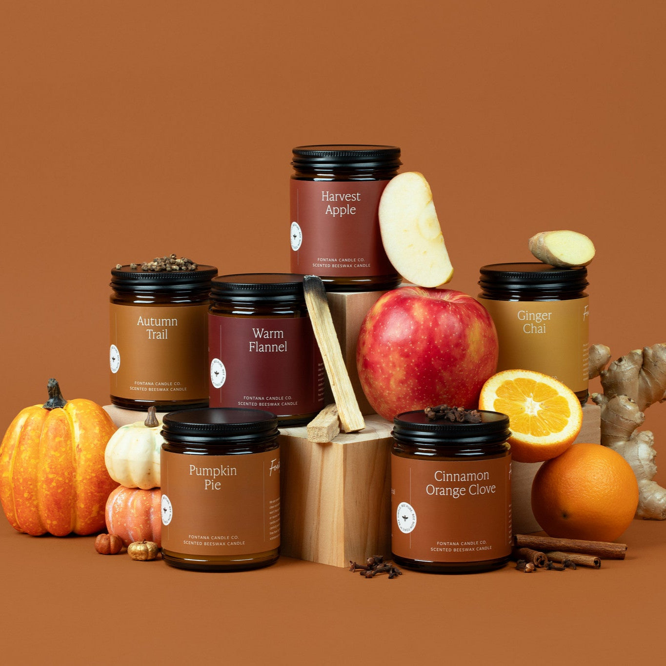 Collection of 6 amber glass jar candles with autumn-themed packaging on a brown background, surrounded by pumpkins, apple slices, spices, and cinnamon sticks.