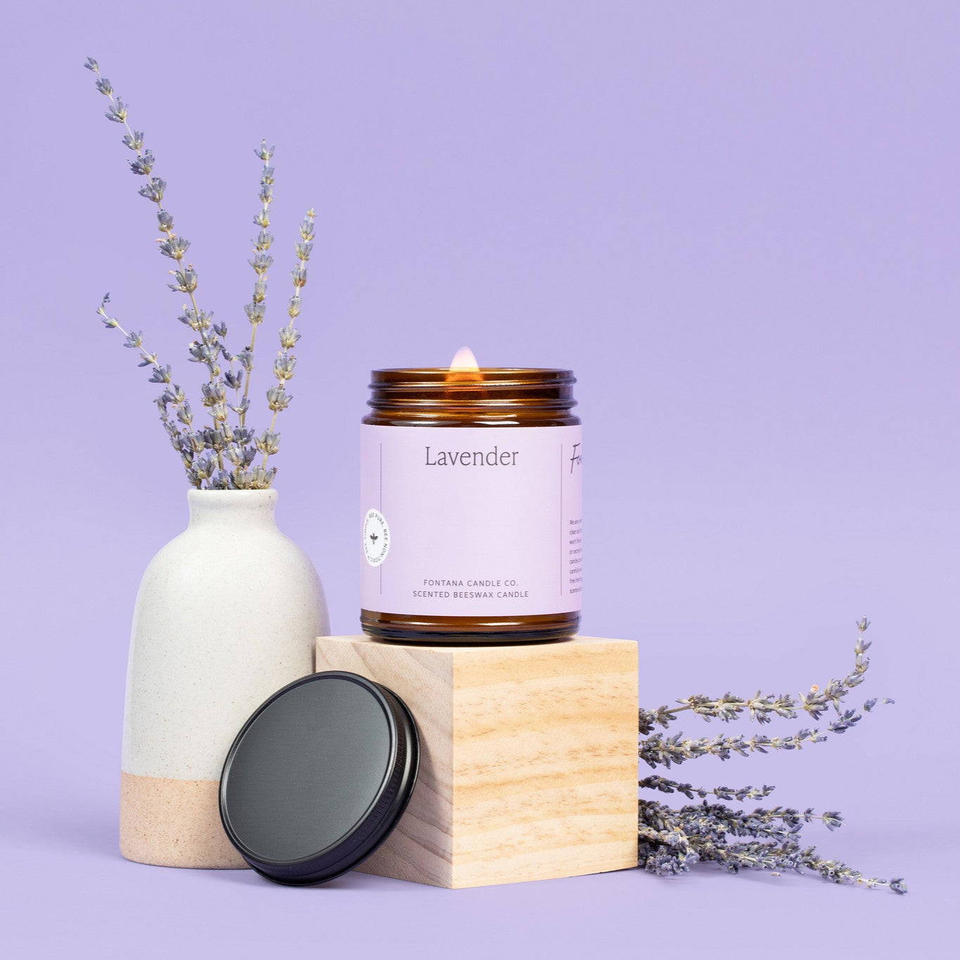 Lit amber glass Lavender jar candle with a light purple label, surrounded by Lavender bunches, in front of a darker purple background. 