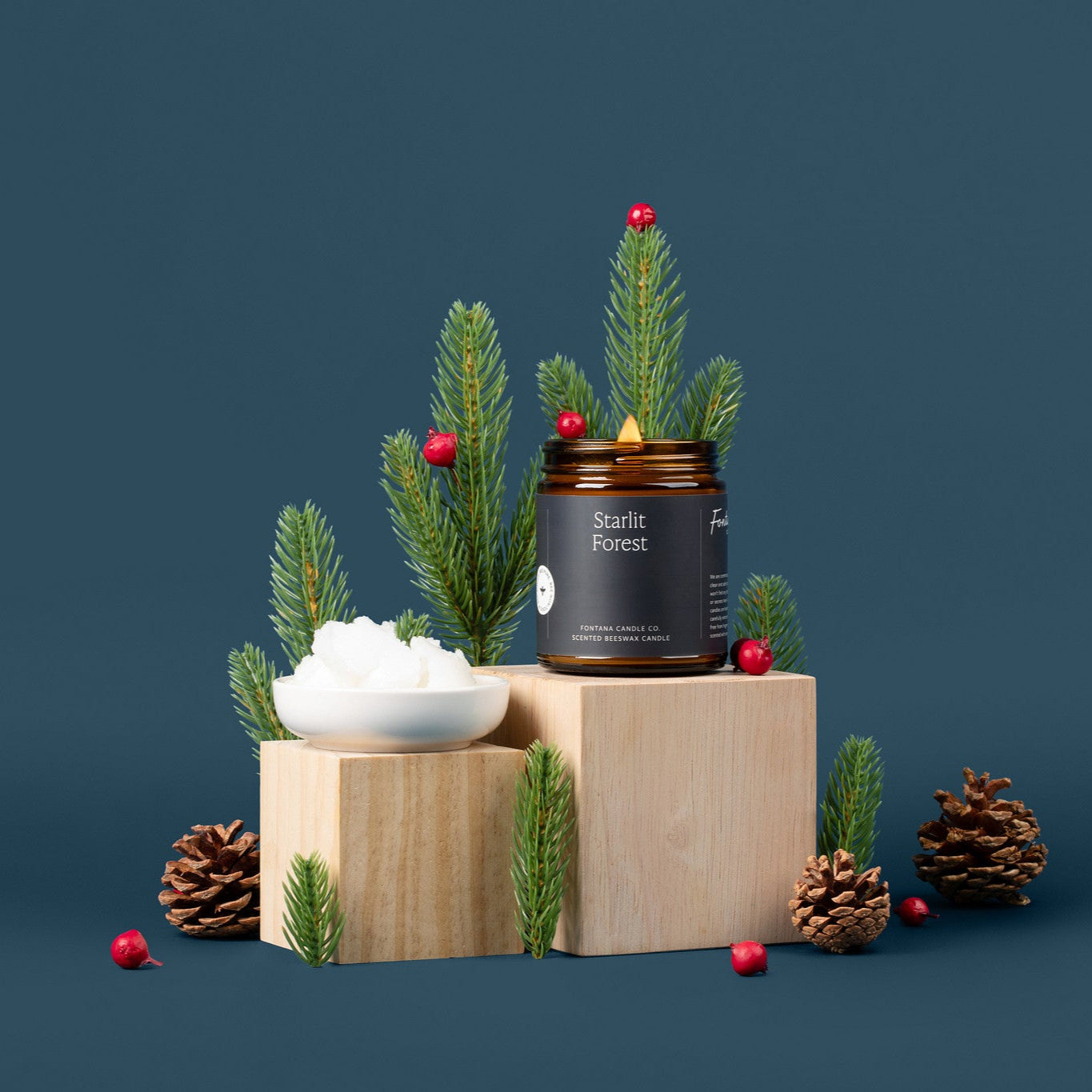 Amber glass jar candle being lit with a black lighter, surrounded by evergreen branches, pinecones, and berries.