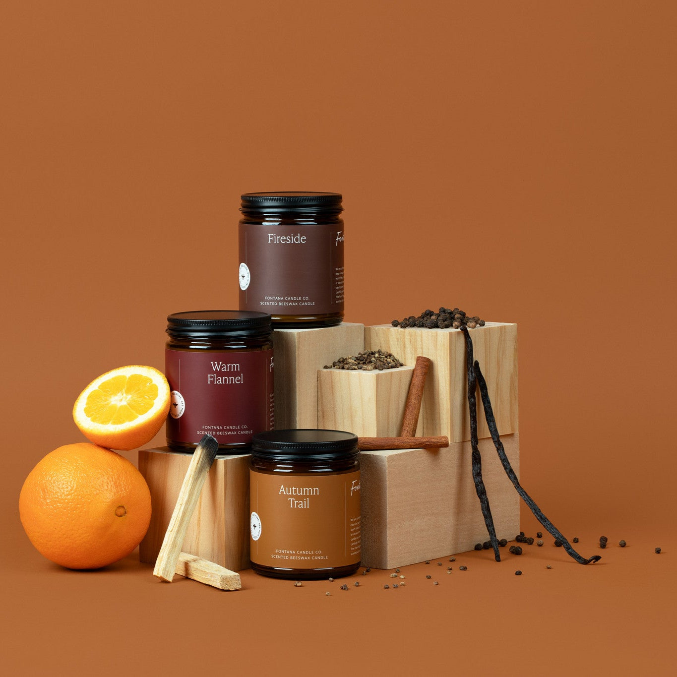 3 amber glass jar candles on a brown background surrounded by oranges, palo santo sticks, cinnamon sticks, and vanilla beans.