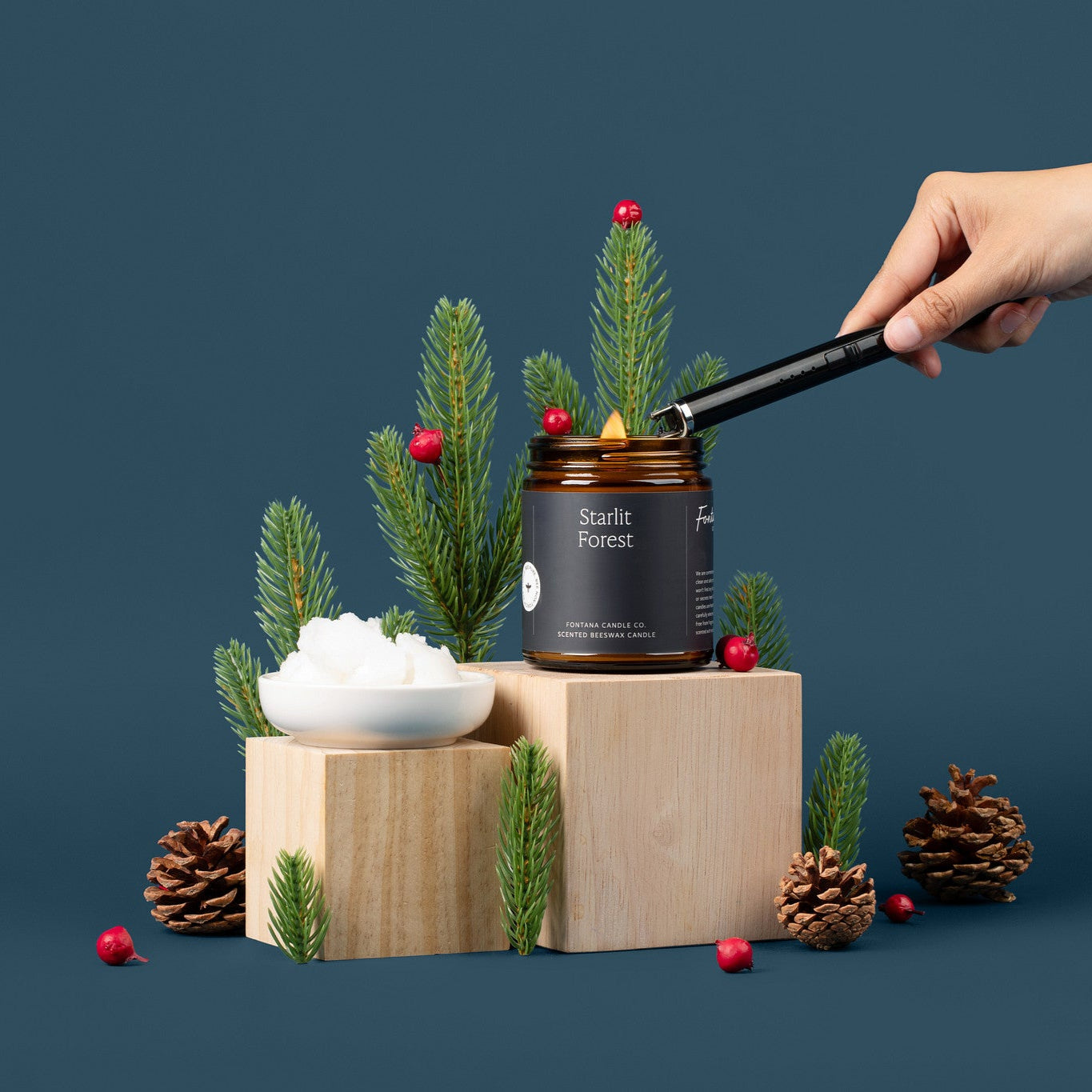 Amber glass jar candle being lit with a black lighter, surrounded by evergreen branches, pinecones, and berries.