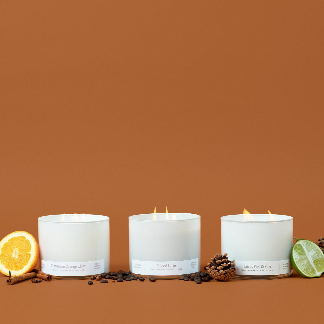 Three lit white glass 3-wick candles on a brown background surrounded by an orange peel, cinnamon sticks, coffee beans, pinecones, and lime.