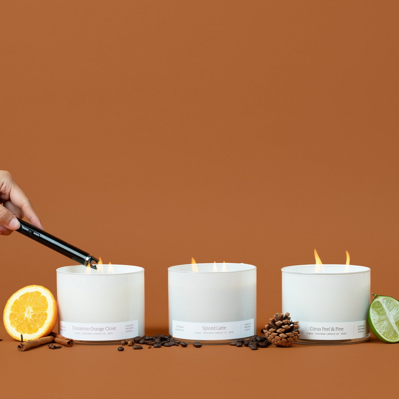 Three lit white glass 3-wick candles on a brown background surrounded by an orange peel, cinnamon sticks, coffee beans, pinecones, and lime.