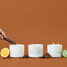 Three lit white glass 3-wick candles on a brown background surrounded by an orange peel, cinnamon sticks, coffee beans, pinecones, and lime.