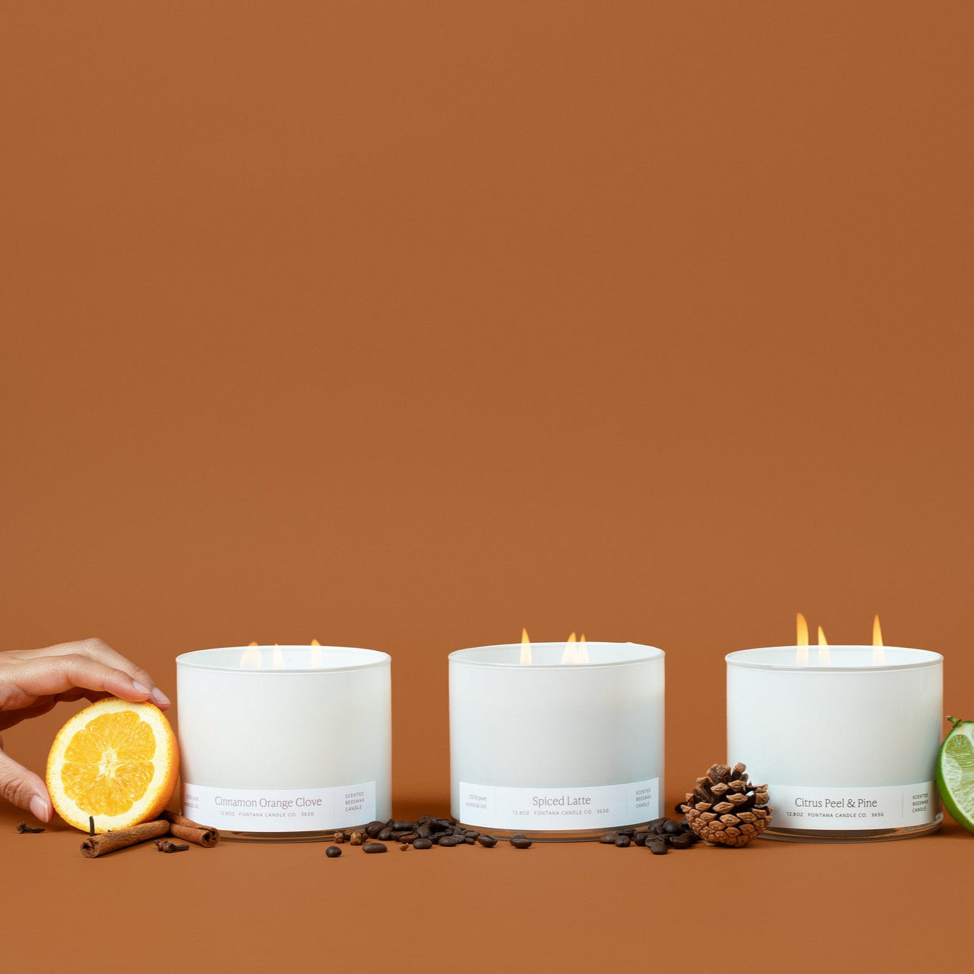 Three lit white glass 3-wick candles on a brown background surrounded by an orange peel, cinnamon sticks, coffee beans, pinecones, and lime.