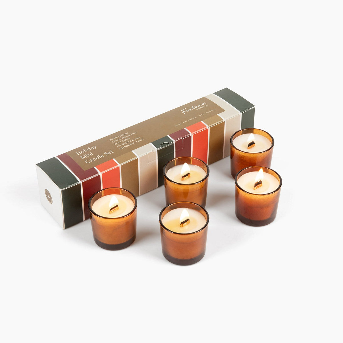 Five lit mini amber glass candles next to a festive holiday box with green, red, and gold, in front of a white background.