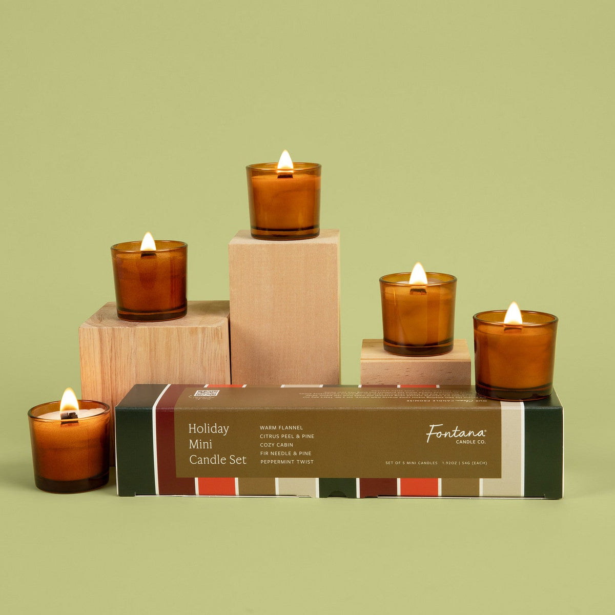 Five lit mini amber glass candles next to a festive holiday box with green, red, and gold, in front of a light green background.