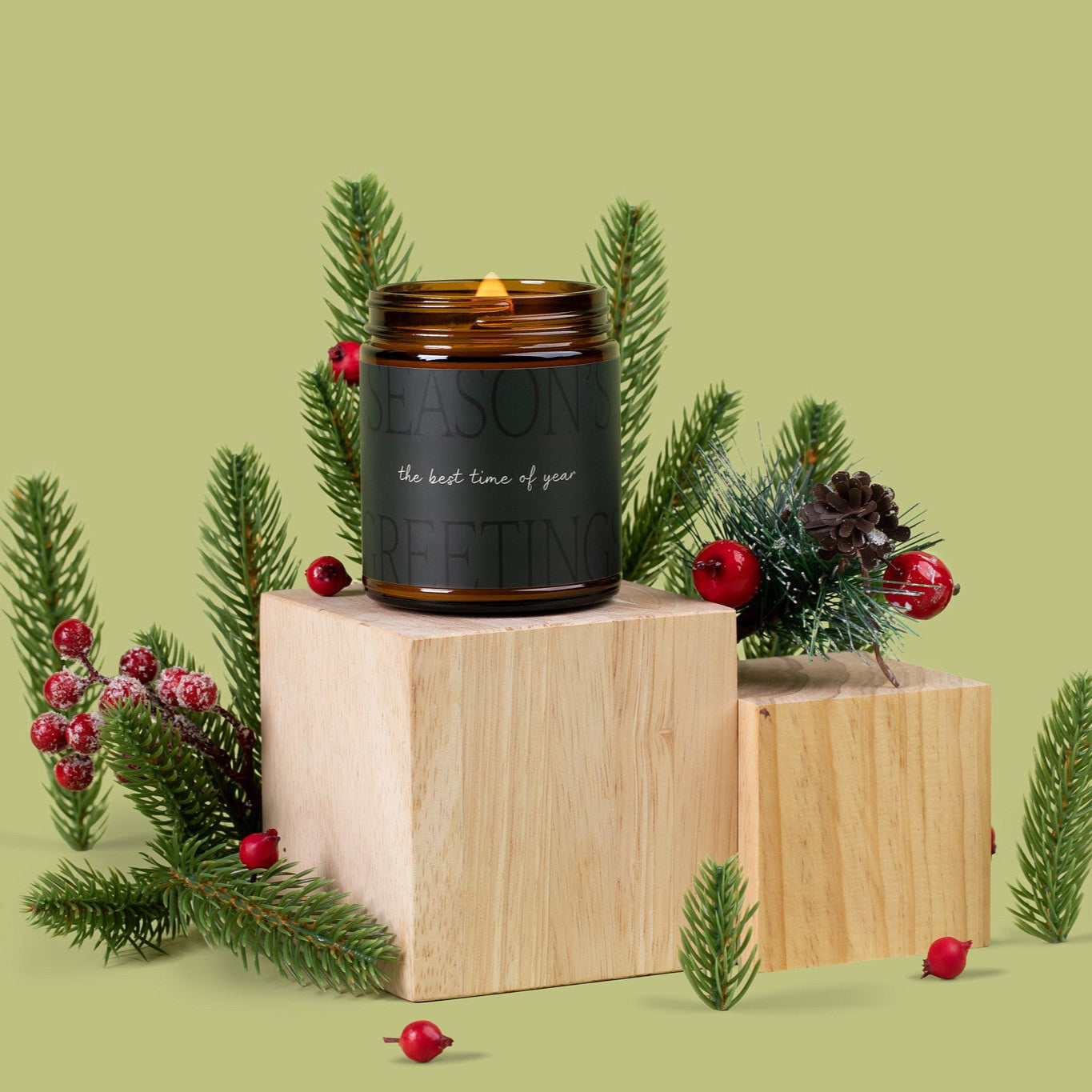 Lit amber glass jar candle with a dark green label that says: "Season's Greetings: the best time of year" on a wooden block with Christmas decorations on a green background.