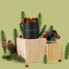 Lit amber glass jar candle with a dark green label that says: "Season's Greetings: the best time of year" on a wooden block with Christmas decorations on a green background.