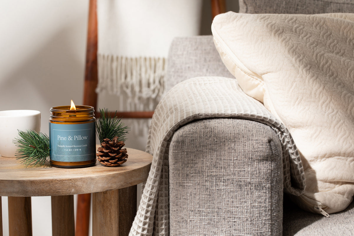 Lit amber glass jar candle on a side table next to a couch with a waffle blanket and pillow. 