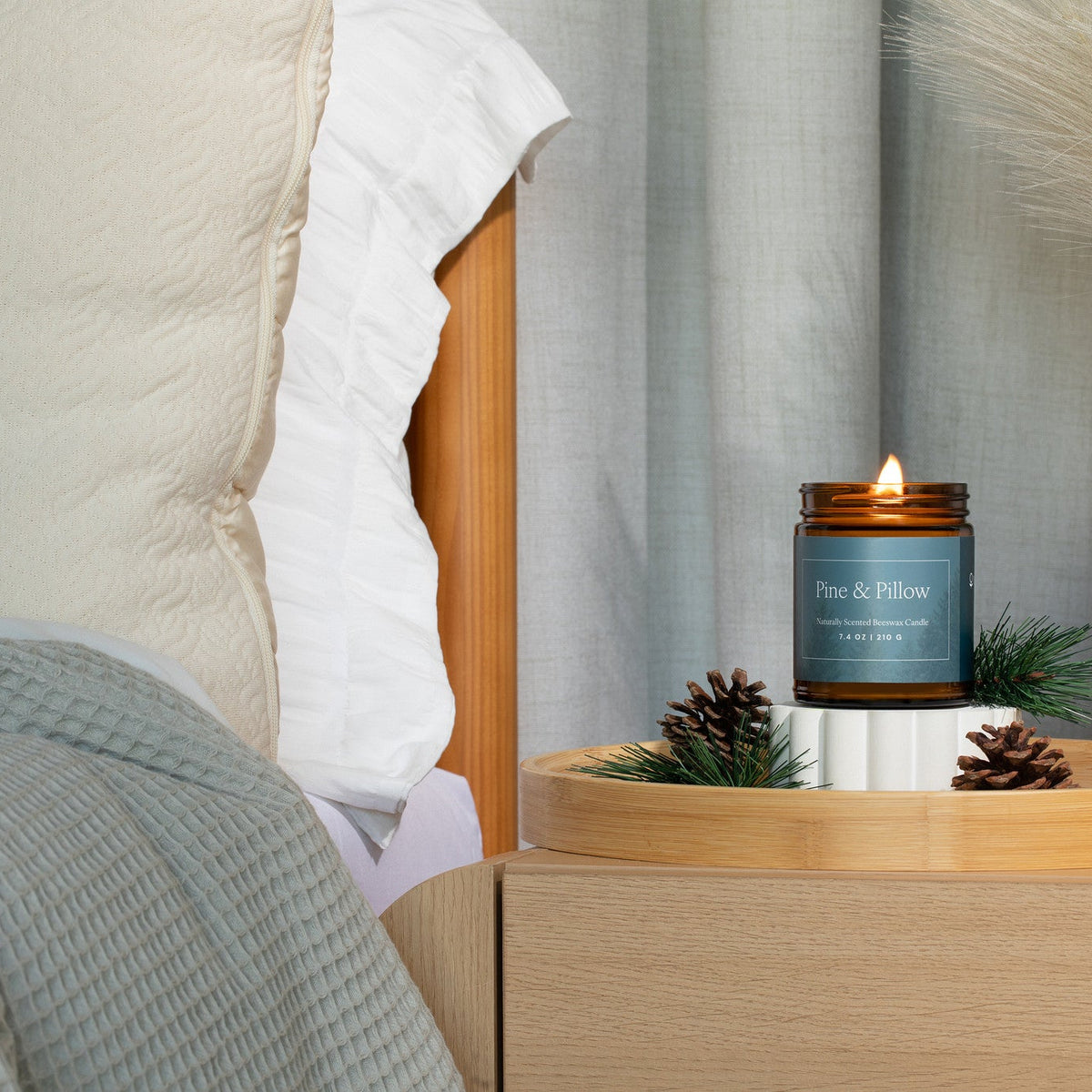 Lit amber glass jar candle on a night stand on a wooden tray, surrounded by pine branches and pinecones, next to a bed. 