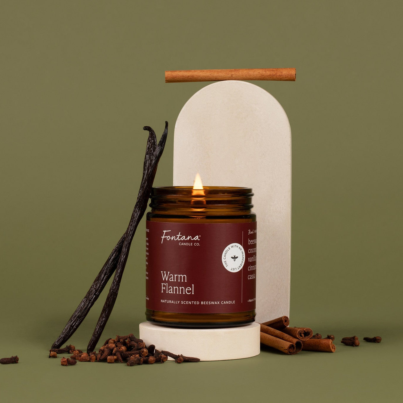 A lit amber glass jar candle with a burgendy red label, in front of a green background, and surrounded by vanilla beans, cinnamon sticks, and cloves.