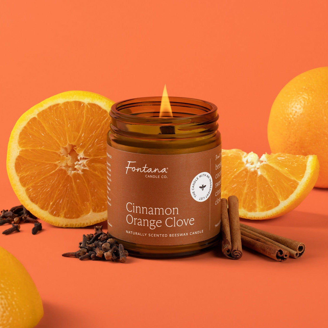 A lit amber glass jar candle labeled Fontana Candle Co. Cinnamon Orange Clove, styled with whole and sliced oranges, cloves, and cinnamon sticks on a solid orange background.