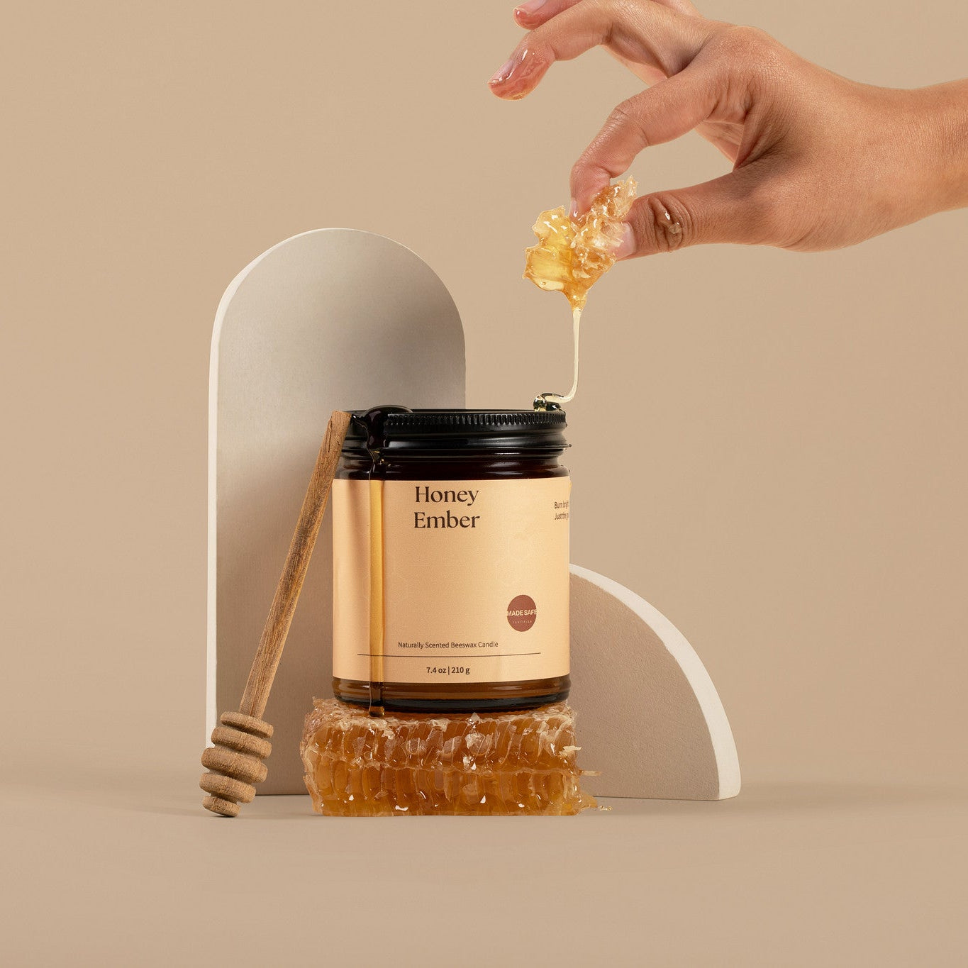 Hand holding honey above a jar candle called Honey Ember with a honeycomb on a beige background