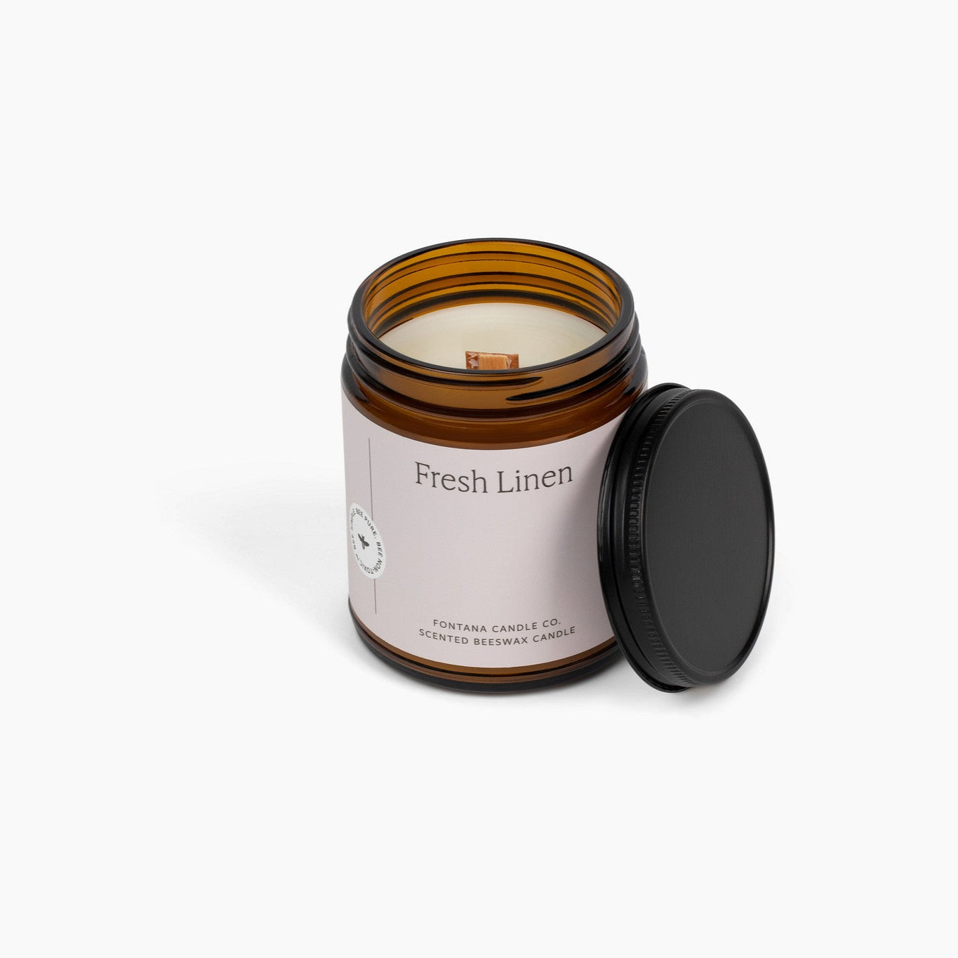 Unlit wooden wick candle in an amber glass jar. The label is a soft pink, and the candle is in front of a white background.