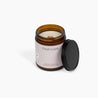 Unlit wooden wick candle in an amber glass jar. The label is a soft pink, and the candle is in front of a white background.