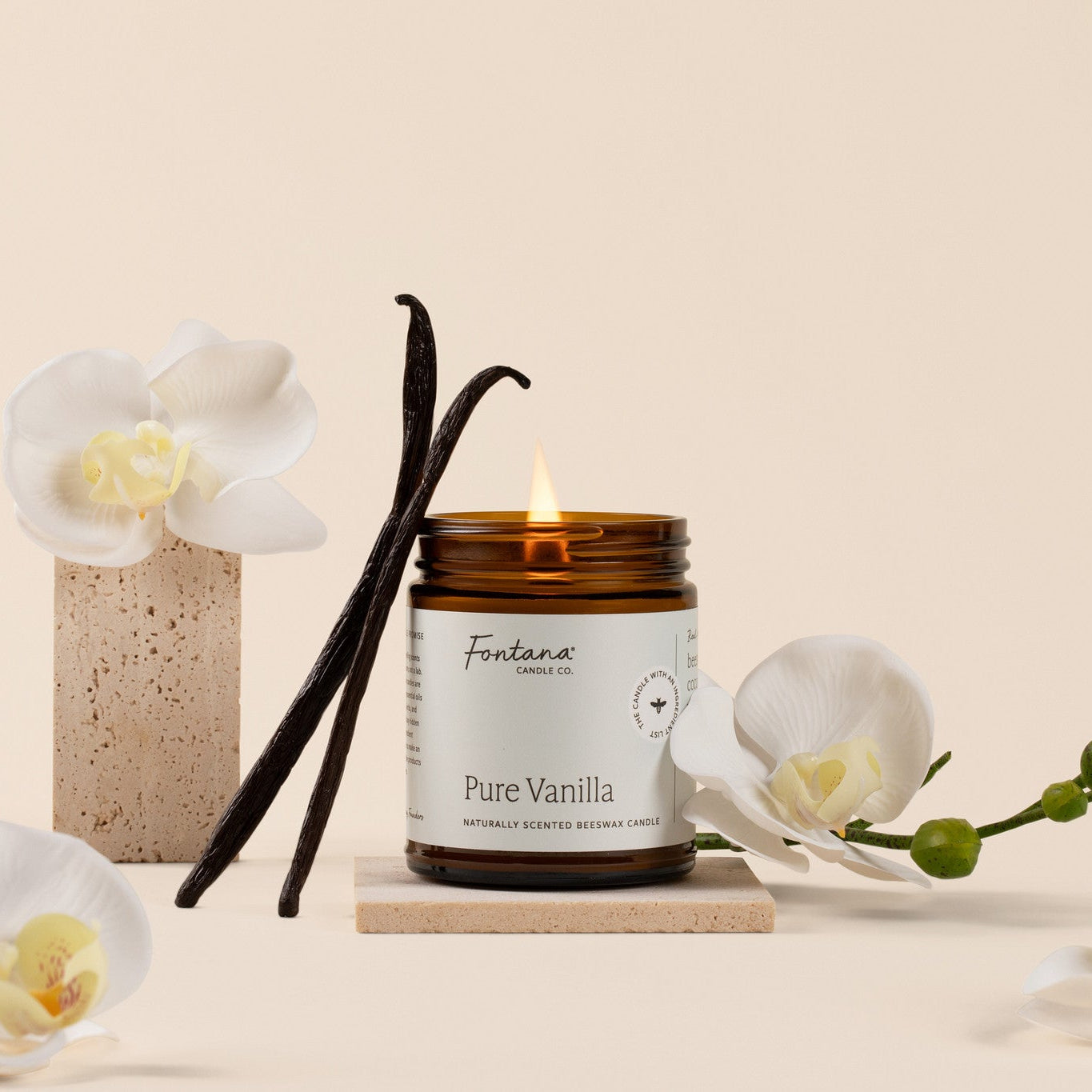 A lit candle in an amber glass jar labeled 'Fontana Candle Co. Pure Vanilla' sits on a small wooden block, styled with vanilla beans, white orchid flowers, and a speckled ceramic vase against a warm beige background.