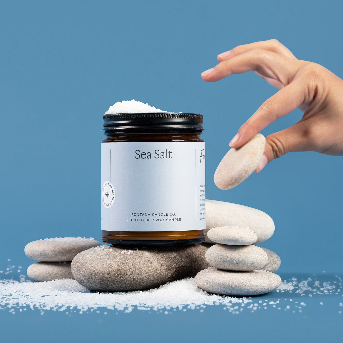 Hand reaching for sea salt over a amber glass jar candle labeled 'Sea Salt' on stones with a blue background.