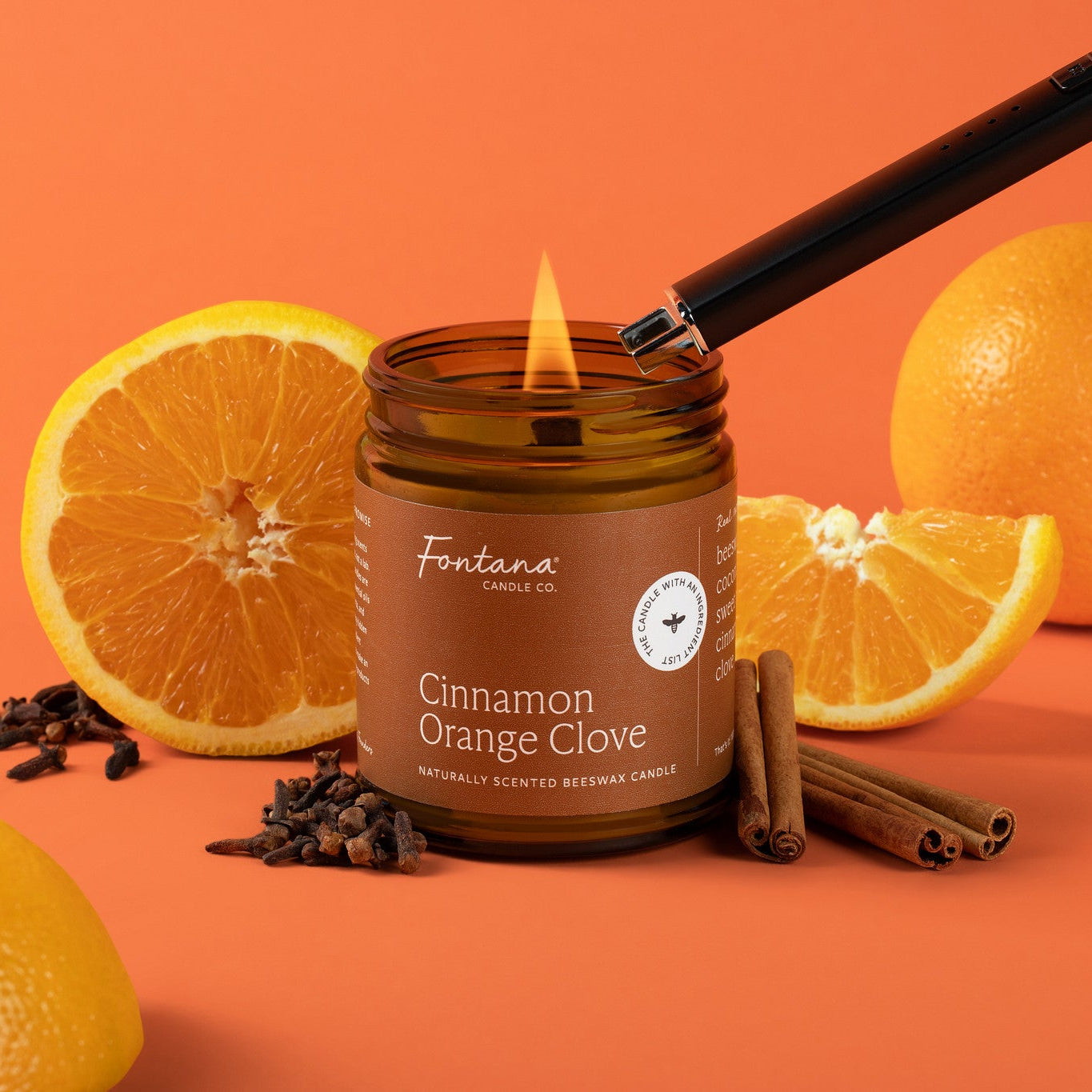 A lit brown glass candle jar labeled Fontana Cinnamon Orange Clove, surrounded by oranges, cloves, cinnamon sticks, and a hand holding a long lighter against a solid orange background.