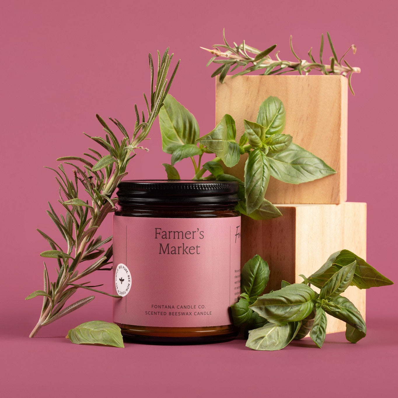Amber glass jar candle labeled 'Farmer's Market' with greenery on a pink background.