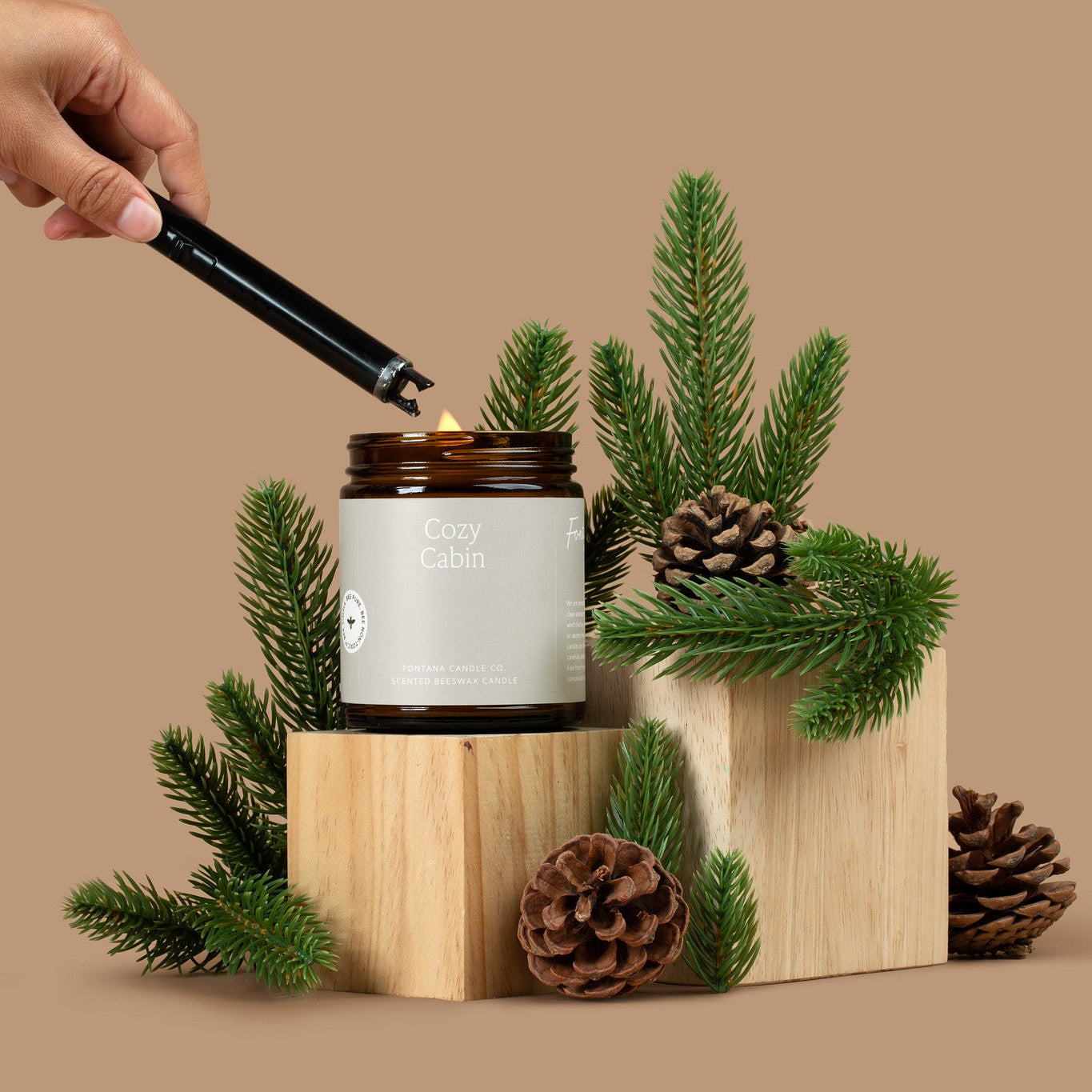 Amber glass jar candle being lit with a black lighter and pine branches on a beige background.