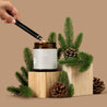 Amber glass jar candle being lit with a black lighter and pine branches on a beige background.